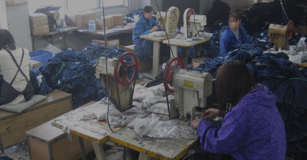 A imagem mostra o ambiente de uma fábrica têxtil com três mulheres trabalhando em máquinas de costura. O espaço é precário e há pilhas de calças jeans espalhadas.