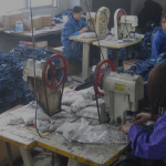 A imagem mostra o ambiente de uma fábrica têxtil com três mulheres trabalhando em máquinas de costura. O espaço é precário e há pilhas de calças jeans espalhadas.