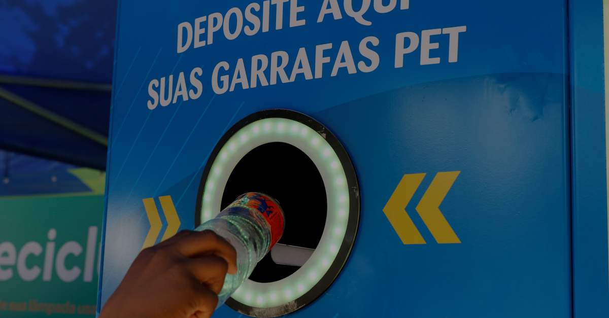 Máquina coletora de resíduos para reciclagem em ação educativa com os dizeres "Deposite aqui suas garrafas PET" e "Insira uma garrafa por vez". Uma garrafa PET está sendo depositada.