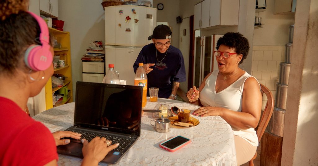 Mulher com fone de ouvido trabalha ao notebook em uma cozinha de uma casa. No mesmo ambiente há mais duas pessoas, um homem e uma mulher