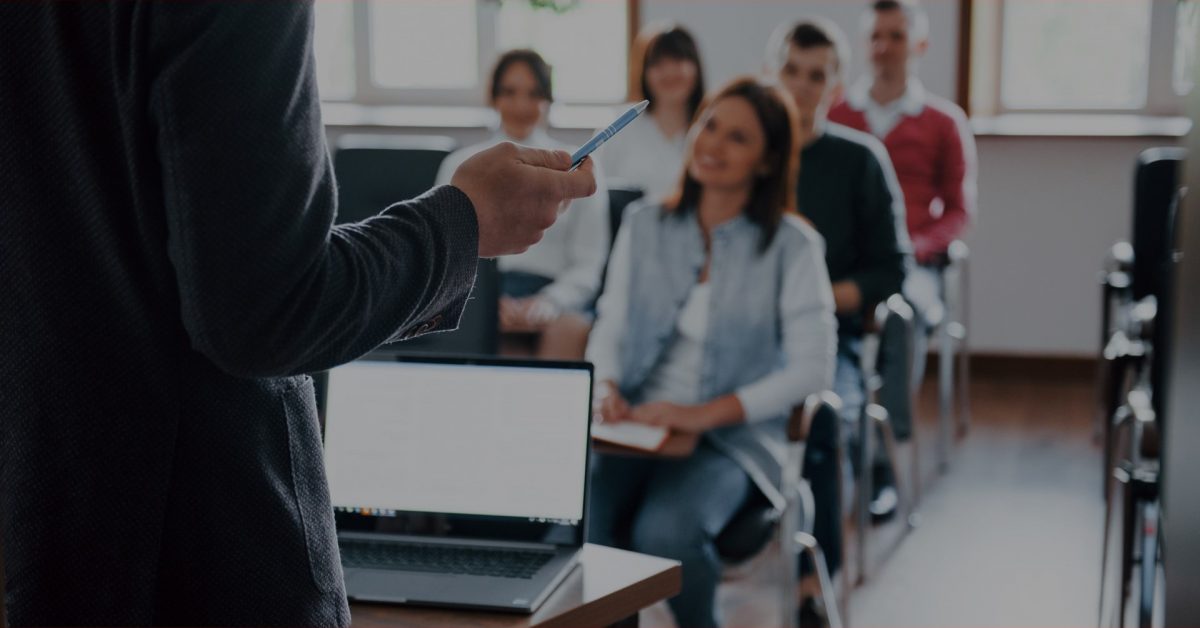 Aula sendo ministrada em sala de aula com estudantes ao fundo