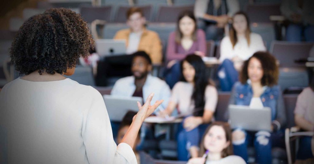 Profissional lecionando de costas em primeiro plano e alunos desfocados em segundo plano