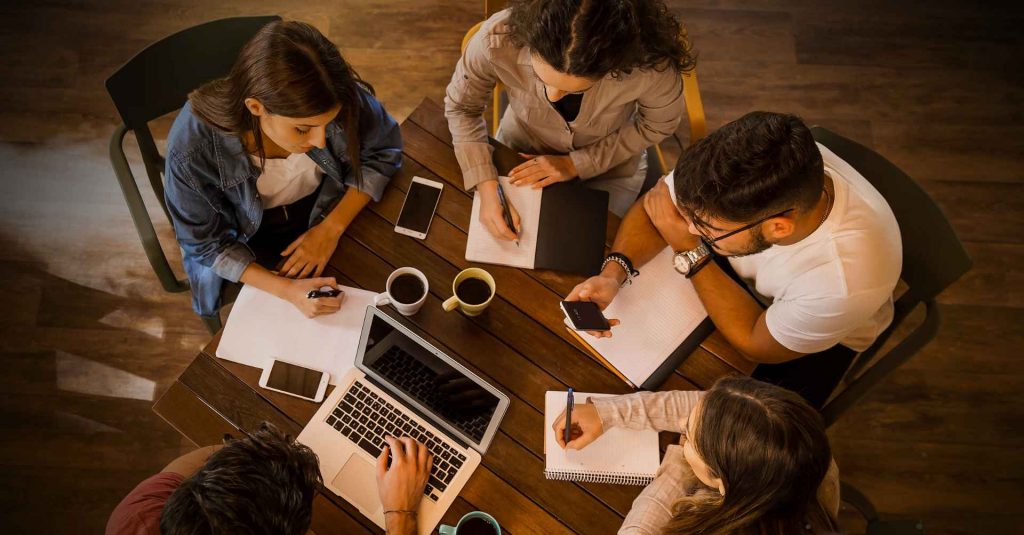 Vista superior de pessoas estudando coletivamente, com computadores, livros e cadernos sobre a mesa